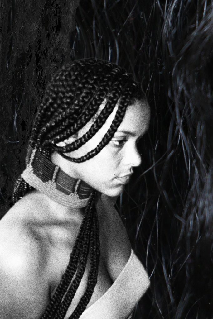 A dark-skinned person with long black braids is shown in profile. They are wearing a tube top and a thick necklace made of beads. They are in front of the same background of black hair.