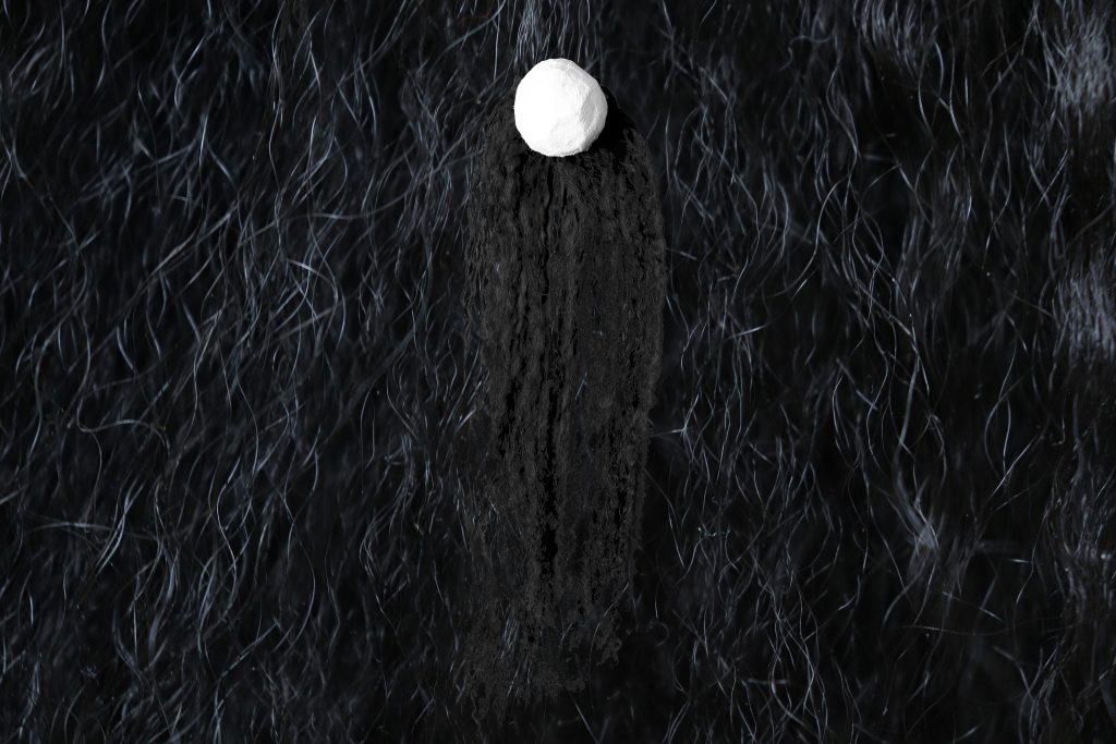 A high-resolution, abstract black and white image of a white ball that seems to be made roughly of clay. Black, textured, crimped hair hangs down from the ball. The hair and ball are on a background of black, long, curly hair highlighted with white.