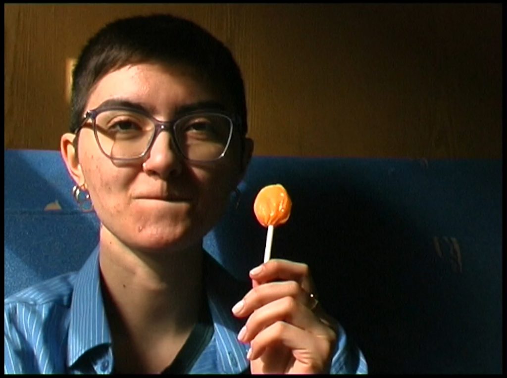 Junge Person mit kurzen dunklen Haaren, Brille und orange-farbenem Lolli in der linken Hand schaut in die Kamera und beißt sich grinsend auf die Unterlippe.
