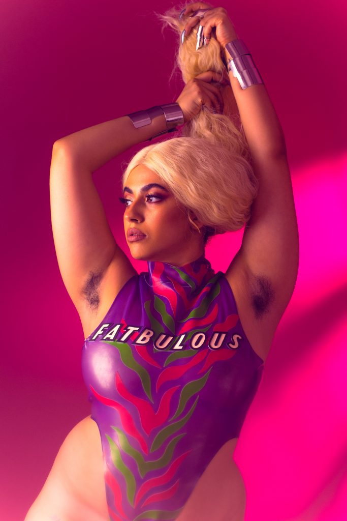 A person with long platinum blond hair stands in front of a brightly pink background. The person is lifting their hair up with both their arms. They are wearing a purple, pink and green coloured bodysuit on the front which it says: "Fatbulous". The model is looking to the left side of the frame. They have dark eyebrows, lashes and eyes.