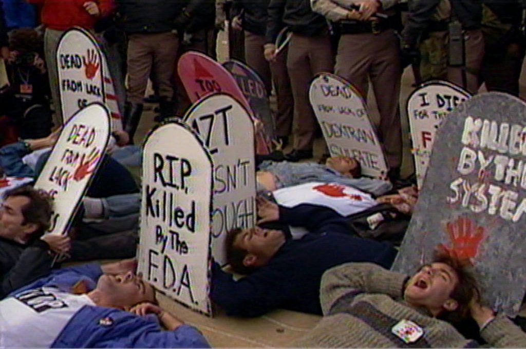 Viele Personen liegen auf dem Boden. Sie halten Kartonschilder in dem Form von GRabsteinen. Text steht darauf "RIP von dem FDA getötet" oder "Ich habe das Placebo bekommen".