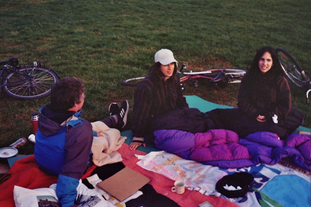 Three people sit, warmly dressed in evening light, on colourful blankets in the park.